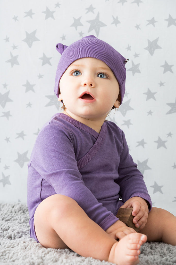 Infant with blue eyes sitting on a grey carpet wearing a purple organic baby hat and kimono